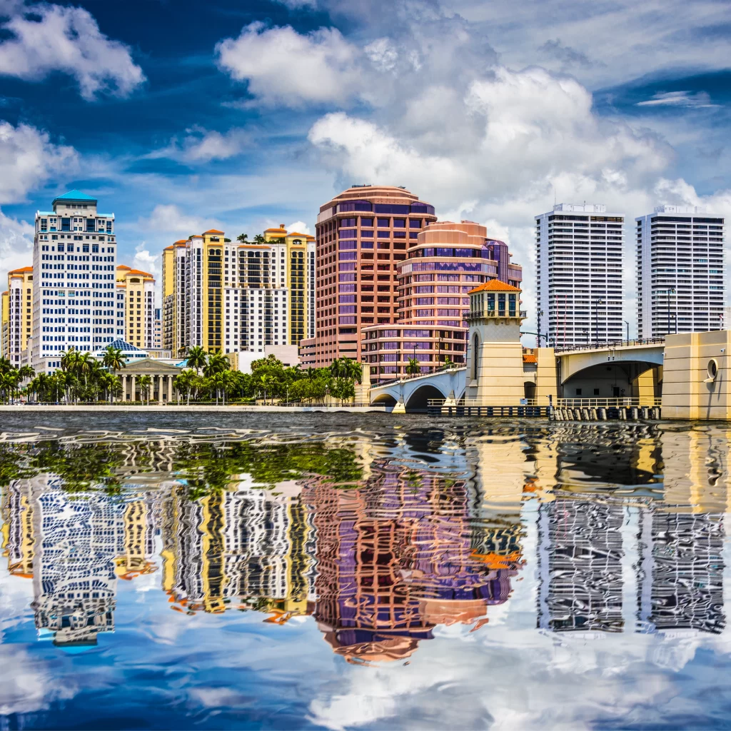 City skyline with several high-rise buildings, a bridge, palm trees, and their reflections on the water under a partly cloudy sky.