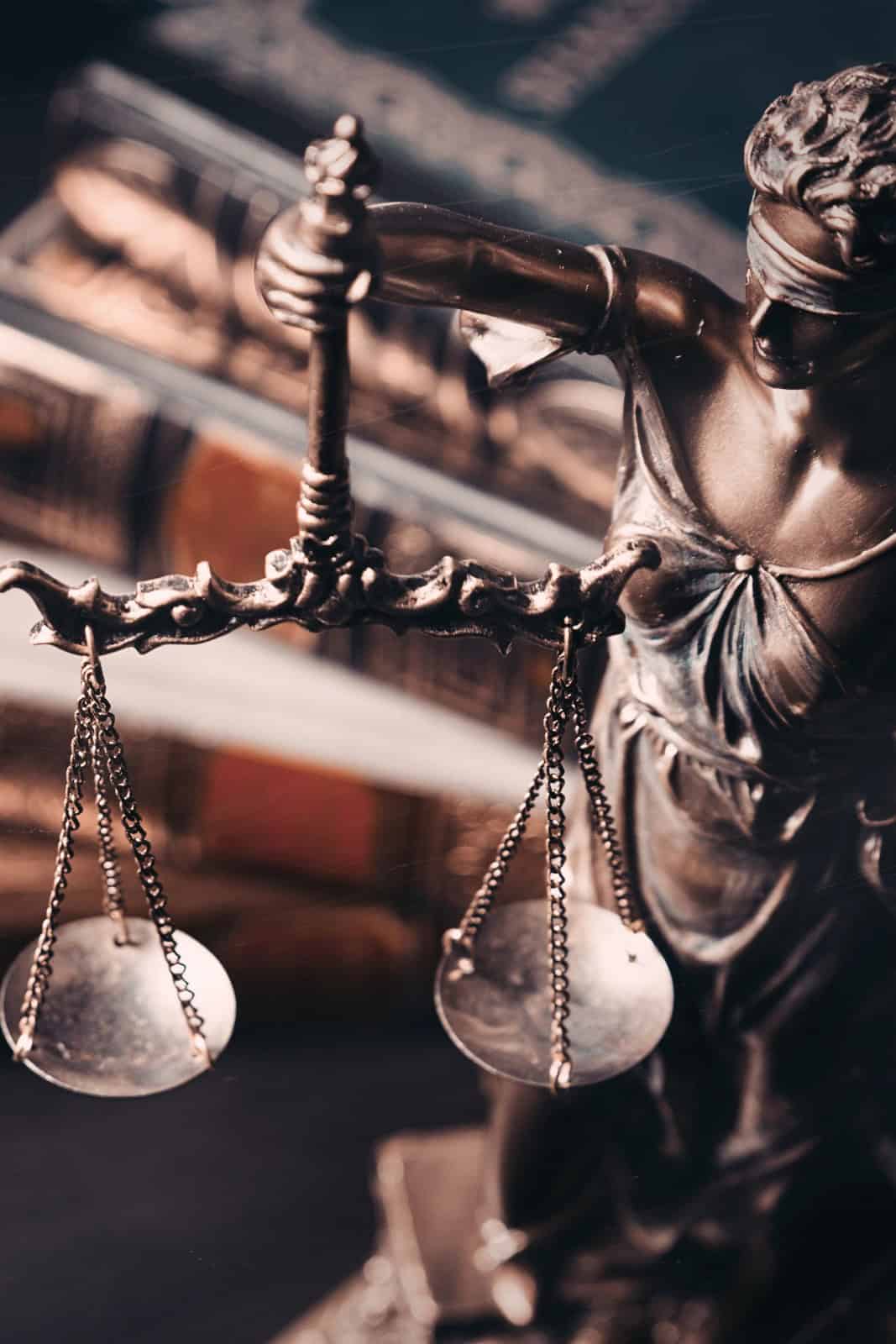 Close-up of a bronze Lady Justice statue holding balancing scales, with law books in the blurred background.