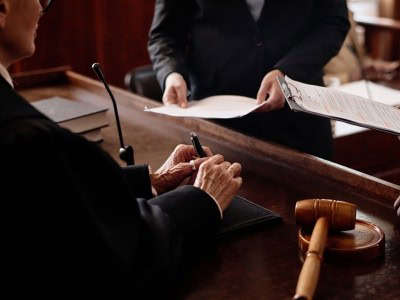 A judge sits at a courtroom bench with a gavel nearby, signing a document while another person hands over paperwork.