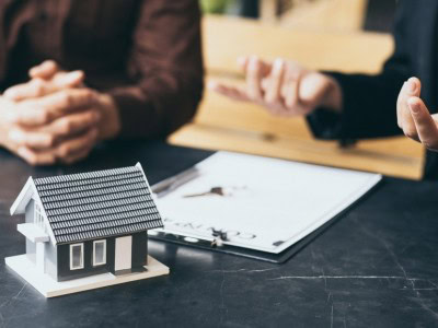 Two people sit at a table with a model house and a clipboard, discussing documents related to real estate or property.