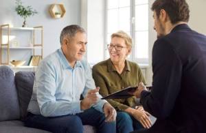 A middle-aged couple sits on a couch talking to a man in a suit who is holding a clipboard in a bright, modern living room.