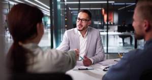 Three people sit at a table in a modern office; a man in a light suit is shaking hands with a woman across the table while another man observes.
