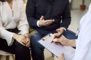 Doctor writing on clipboard while seated couple speaks during medical consultation