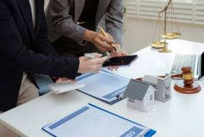 Two people review documents and a smartphone at a desk with a gavel, scale, house model, calculator, and contract paperwork.