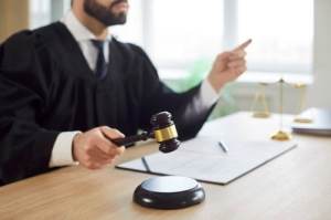 A judge in a black robe holds a gavel and points while sitting at a desk with legal documents and a scale of justice.