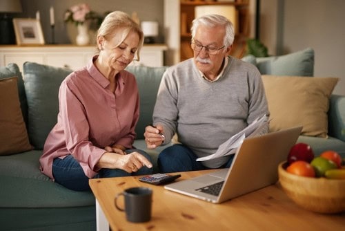 An older man and woman sit on a couch looking at papers and using a calculator, with a laptop and a bowl of fruit on the table in front of them.