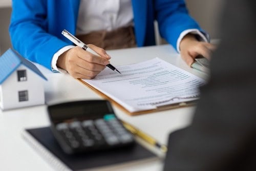 Person in a blue blazer signing documents on a clipboard at a desk with a small house model, calculator, and notebook.