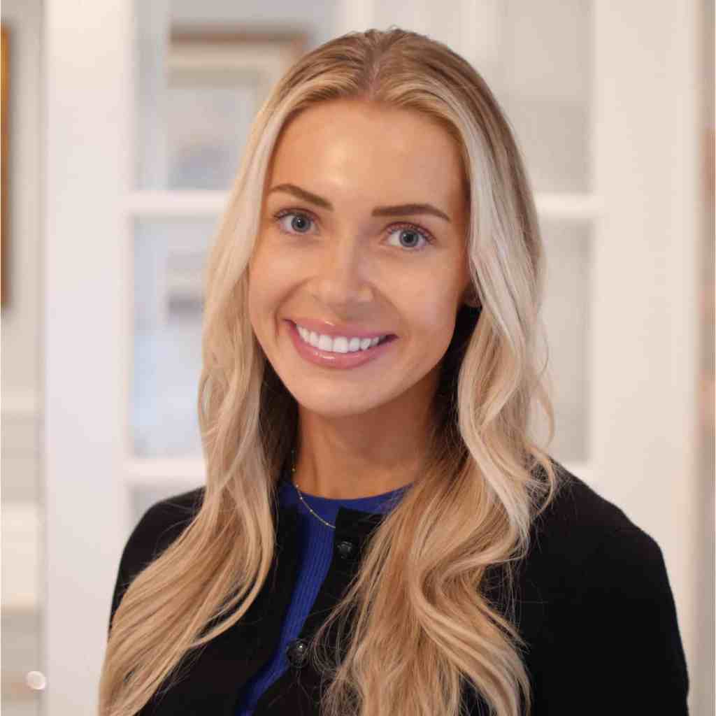 A woman with long blonde hair and a black top smiles at the camera in a well-lit indoor setting with white walls and glass doors in the background.