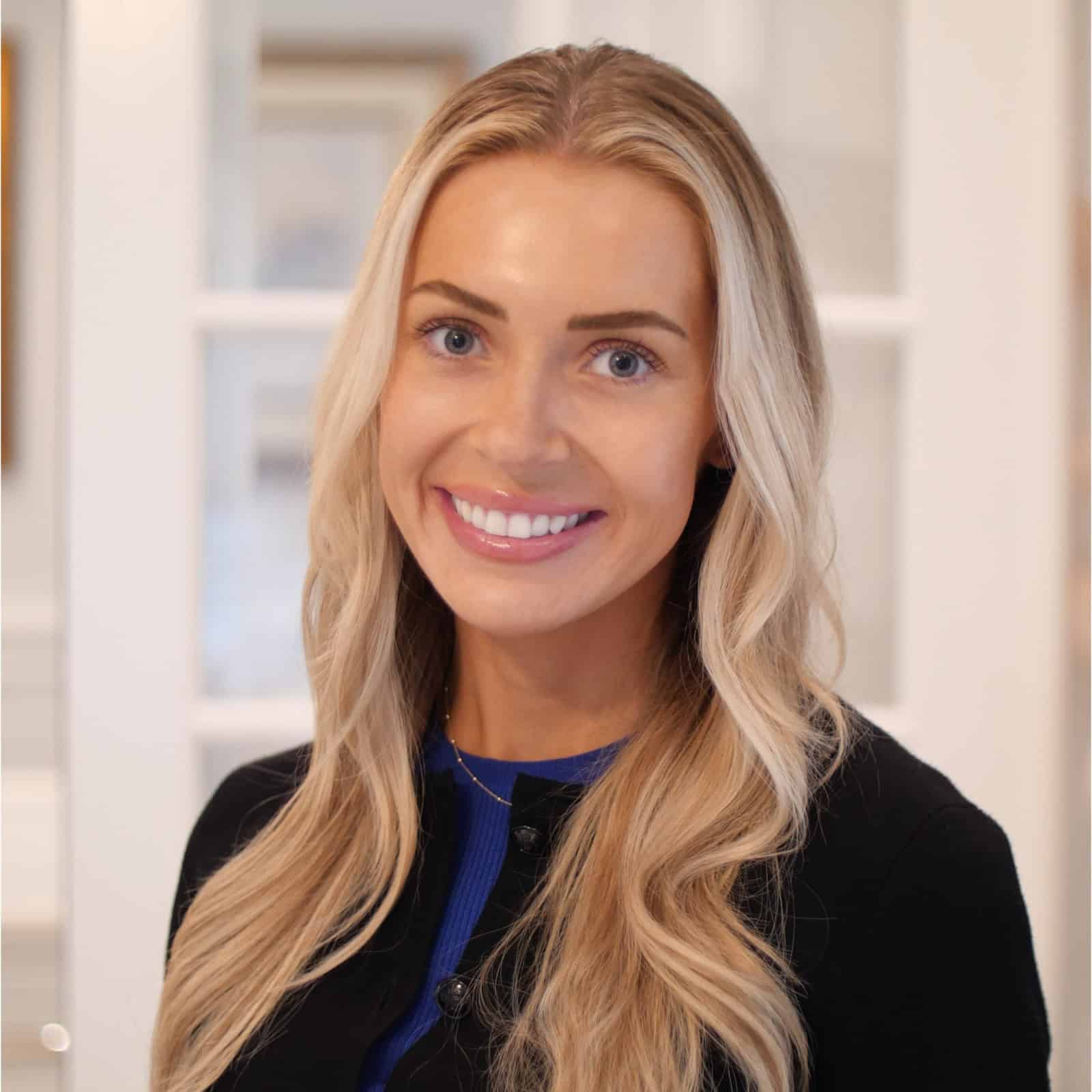 A woman with long blonde hair and a black top smiles at the camera in a well-lit indoor setting with white walls and glass doors in the background.
