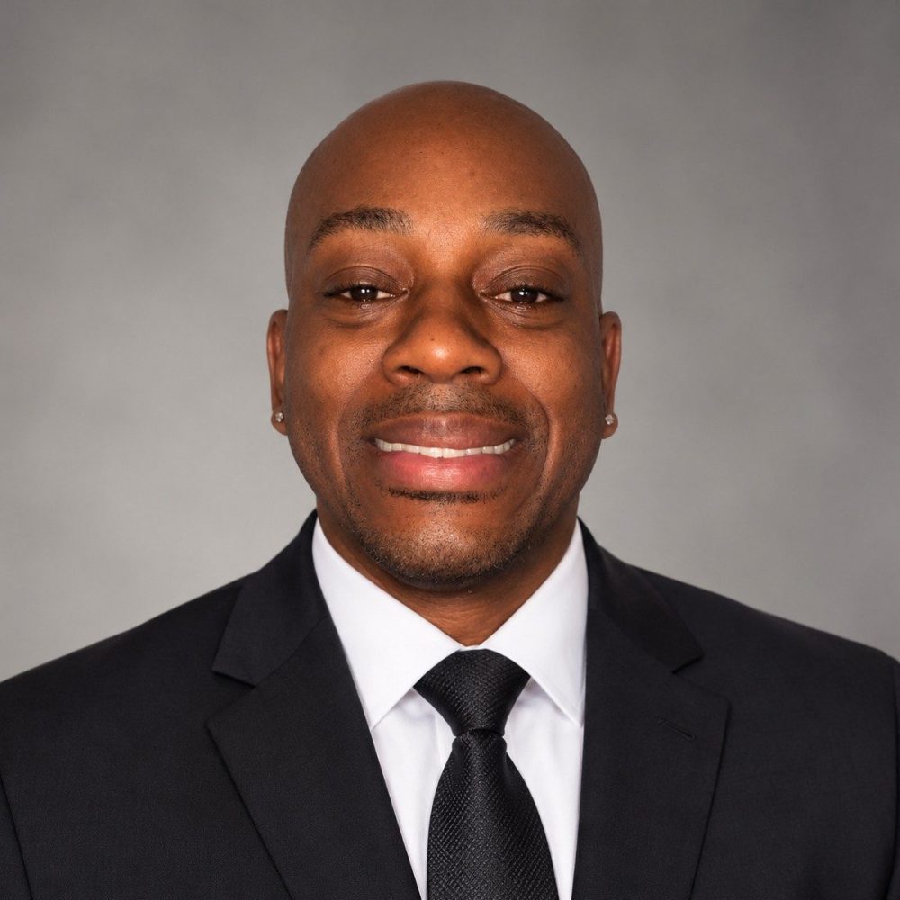A man wearing a black suit, white shirt, and black tie smiles at the camera against a plain gray background.
